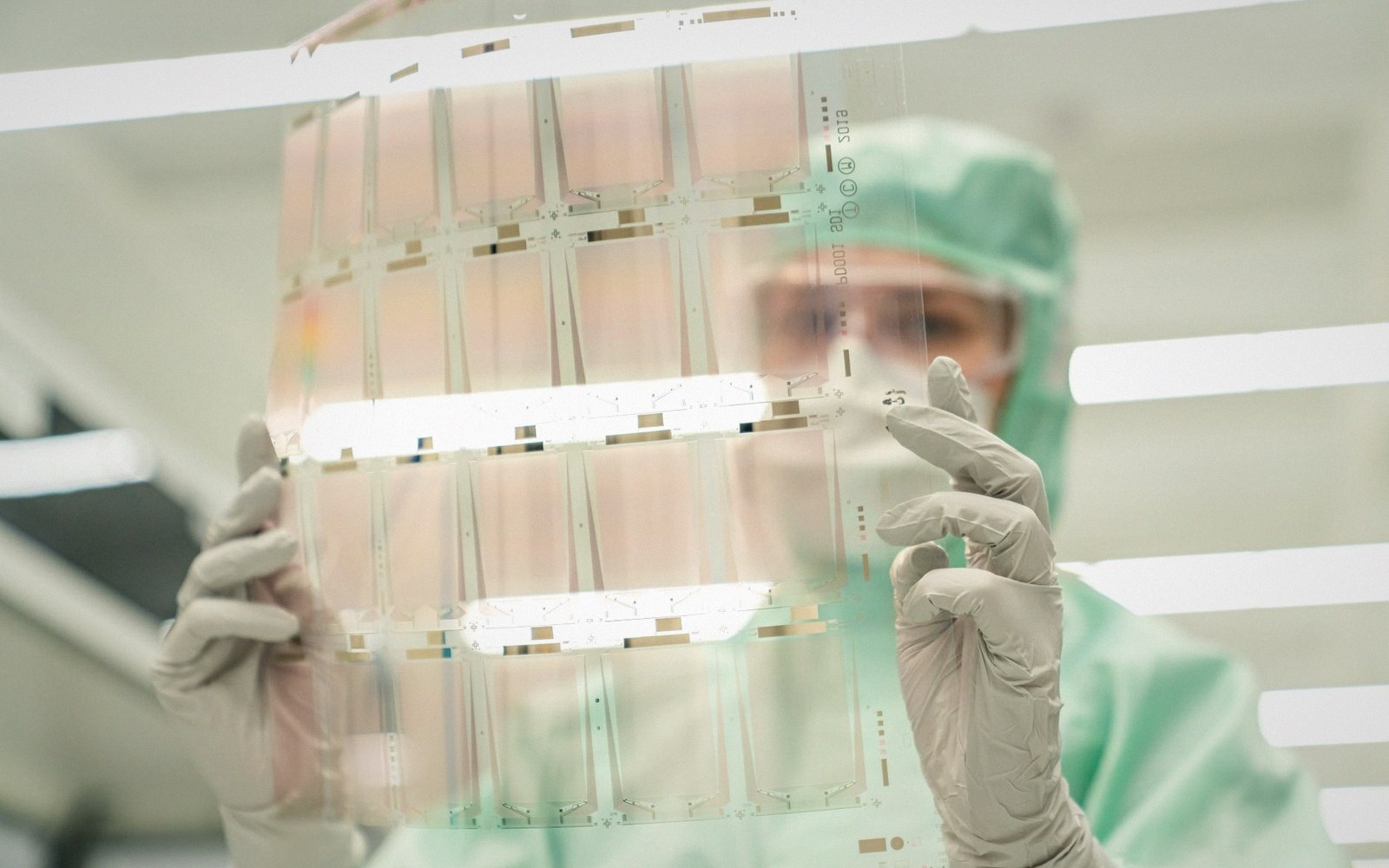 Worker in cleanroom suit examining a thin flexible electronic panel under lab lighting