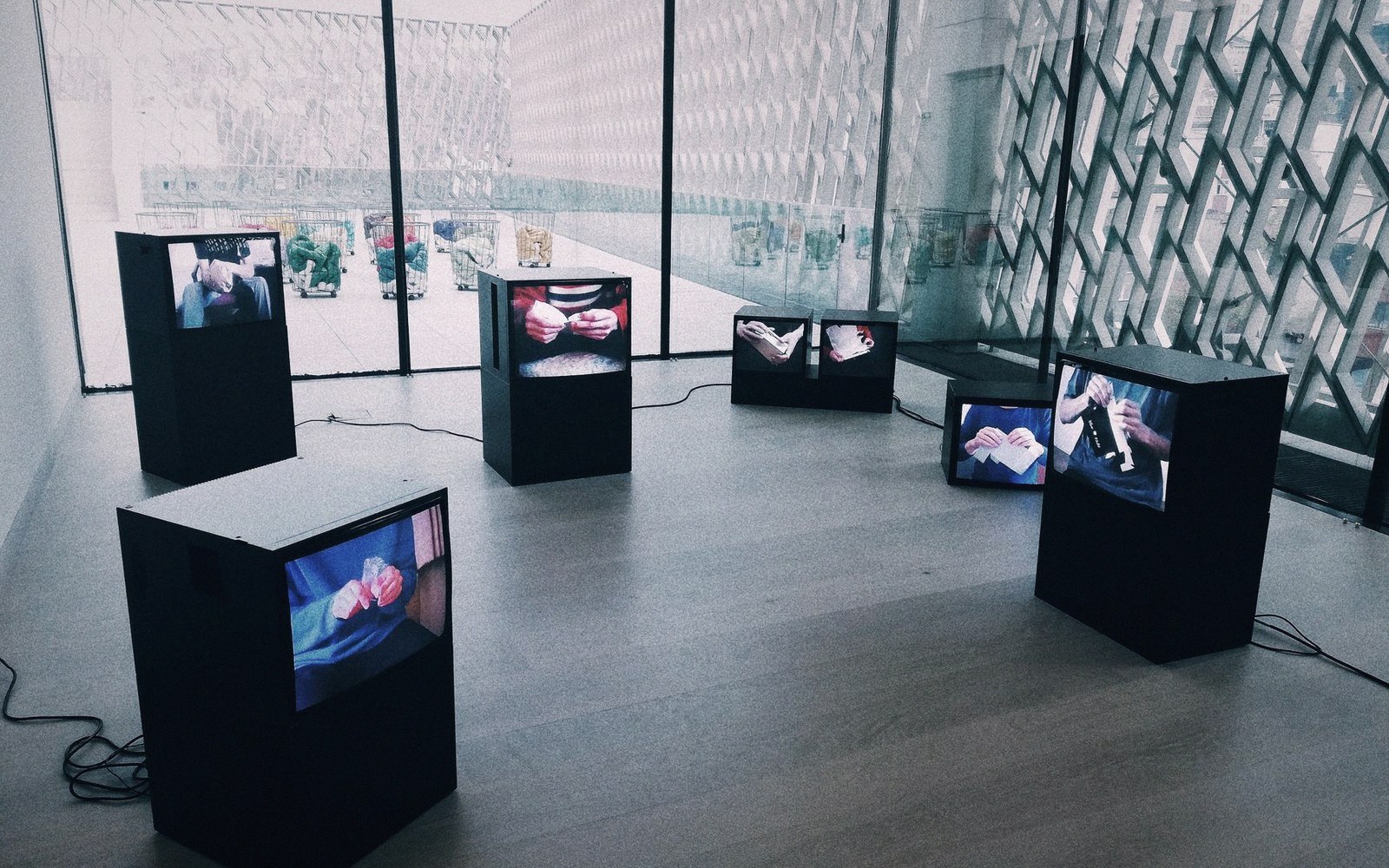 Video art installation with multiple screens on black plinths inside a glass-walled gallery